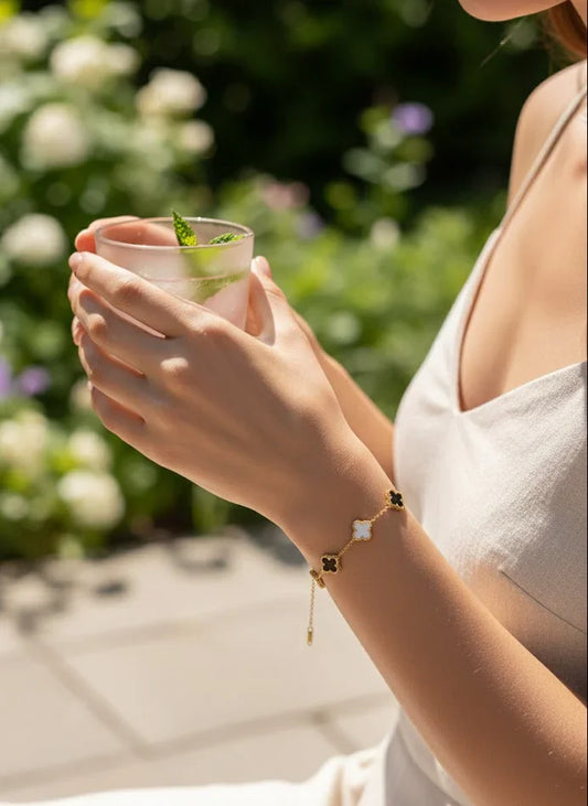Person holding a glass with a drink and a mint leaf, standing outdoors with greenery in the background